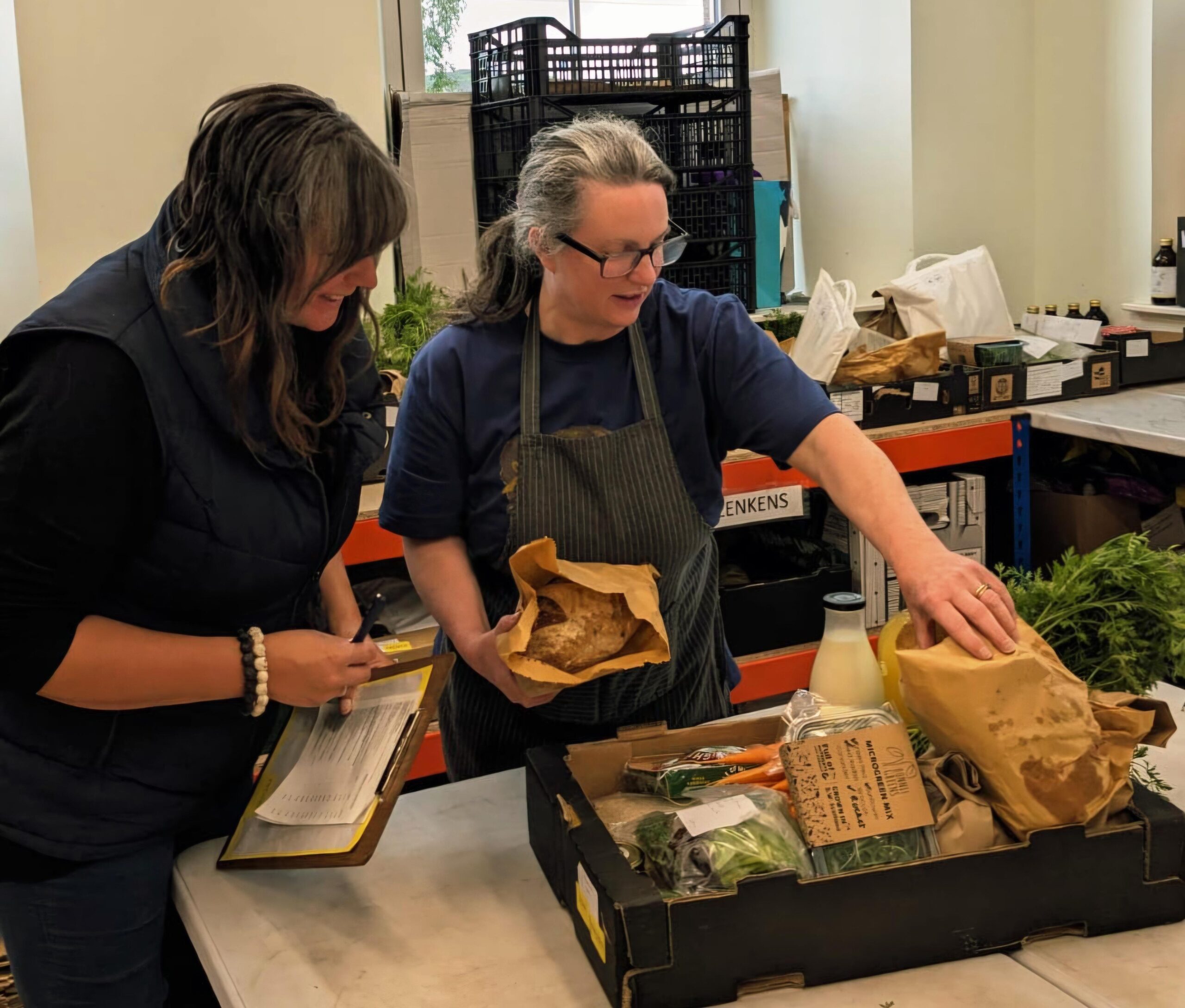 Two women placing food items into a cardboard box ready for delivery